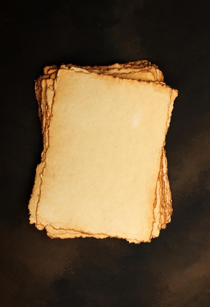 Photo close up stack of several blank old antique vintage brown paper parchment sheets with aged and weathered edges over dark table background