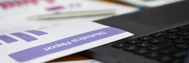 Close-up of statistics report with diagrams and charts laying on office table. Important documents on working desk. Black modern laptop. Business and finance concept