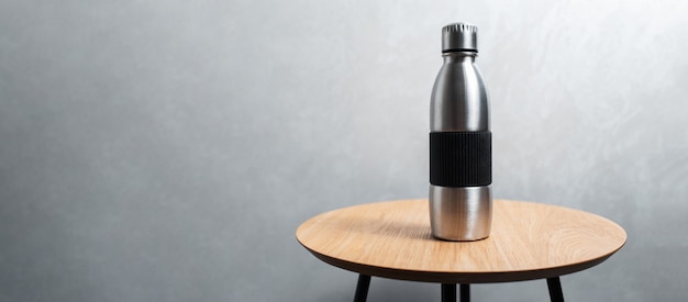 Close-up of steel reusable thermo water bottle on wooden table against background of textured grey wall. Panoramic banner view with copy space.