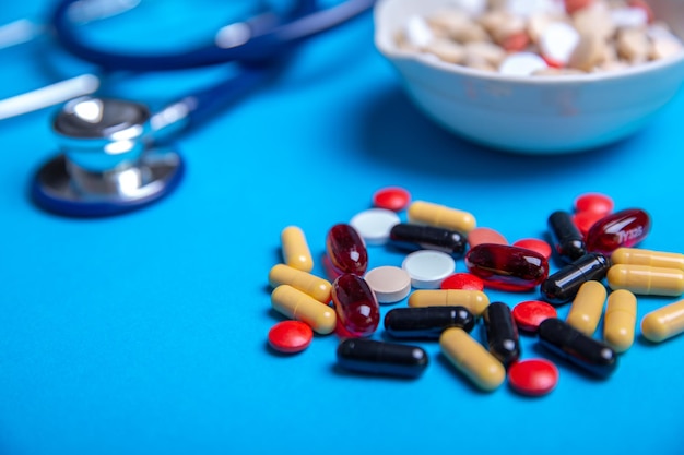 Close up step with pills of several colors in the background. Stethoscope and a bowl with pills. Blue background