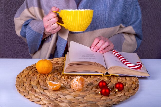 Close-up stilleven met een boek, kerstdecor op tafel. Een vrouw van middelbare leeftijd in een stola houdt een grote gele kop warme drank vast.