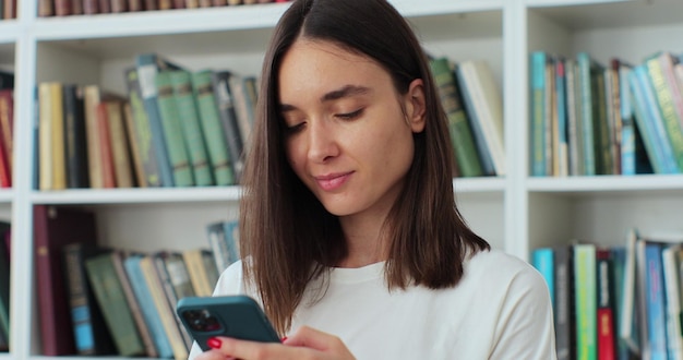 Close up student girl standing in library and using smartphone texting message check online studies timetable using mobile application