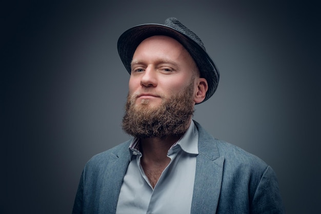 Крупным планом студийный портрет бородатого мужчины в фетровой шляпе на сером фоне.