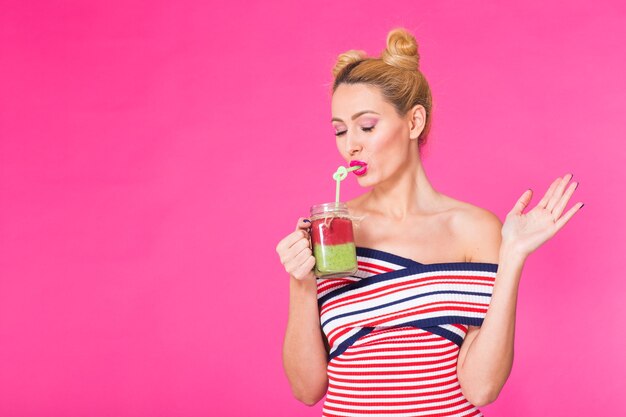 Close-up studio portret van grappige jonge vrouw heldere outfit dragen, houden en lekkere groene smoothie milkshake drinken op roze achtergrond