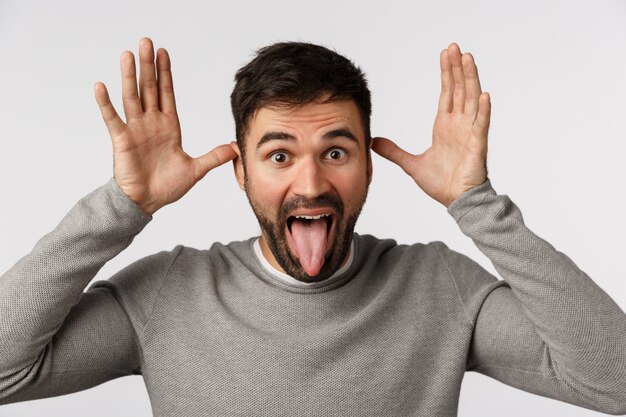 Close-up studio shot carefree and playful, funny cute bearded man in grey sweater, stick tongue and make teasing gesture with hands near ears, fool around, asking catch him playing with kids