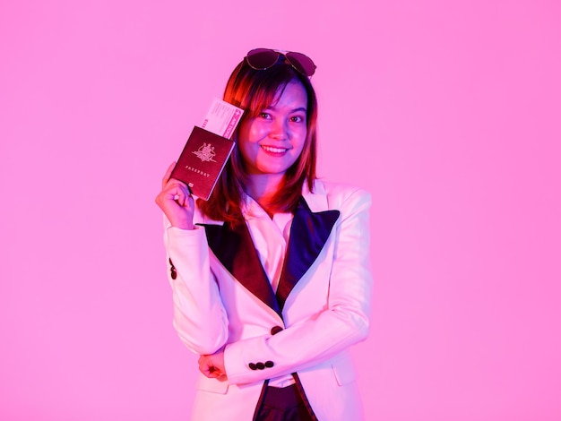 Close up studio shot of traveling passport and airplane ticket in hand of Asian urban rich wealthy female traveler in casual blazer suit and shades look at camera on pink light blurred background.