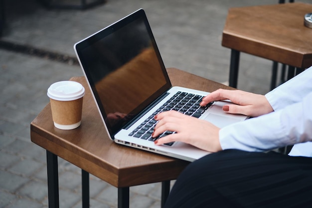 Close up stylish businesswoman with coffee to go working on laptop in cafe on street