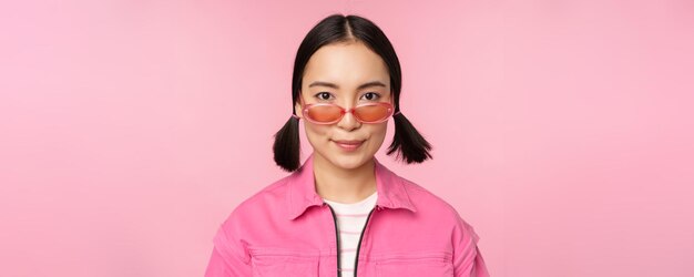 Крупный план стильной корейской девушки в солнцезащитных очках, счастливо улыбающейся, позирующей на розовом фоне.