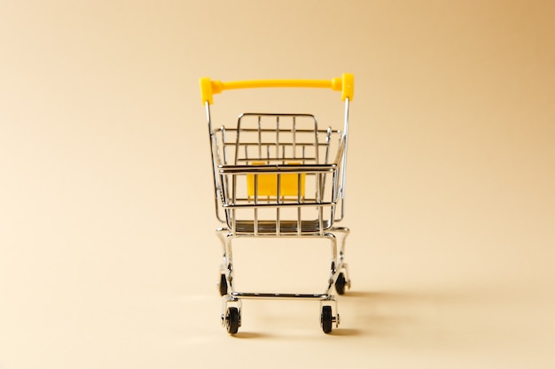 Close up of supermarket grocery push cart for shopping with black wheels and yellow plastic elements on handle isolated on beige background. Concept of shopping. Copy space for advertisement.