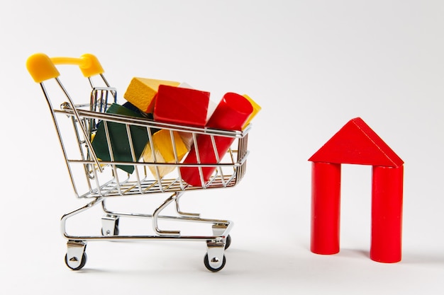 Close up of supermarket grocery push cart for shopping with yellow handle filled with multi-colored geometric shapes isolated on white background. Concept of shopping. Copy space for advertisement.