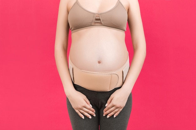 Close up of supporting orthopedic bandage against backache on pregnant woman at pink background with copy space. Orthopedic abdominal support belt concept.