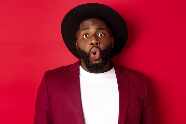 Close-up of surprised black man staring at camera with wonder, standing against red background