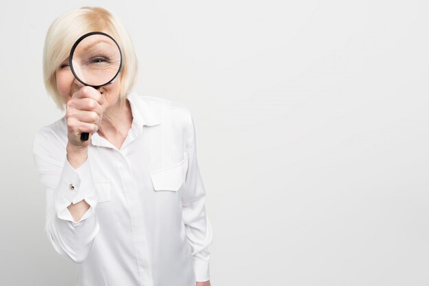 Close up of suspicious old woman in white blouse looking straight ahead through the reading glass. She looks strange. Cut view.