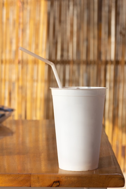 Close up of takeaway blank white disposable cup and tube on wooden table and with a bamboo wall as a background bokeh. summertime.