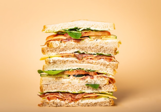 パステルイエローの背景にチーズ、ハム、生ハム、新鮮なレタス、トマト、きゅうりと背の高いカットのおいしいサンドイッチのクローズアップ。朝食やランチにヘルシーなグルメサンドイッチ