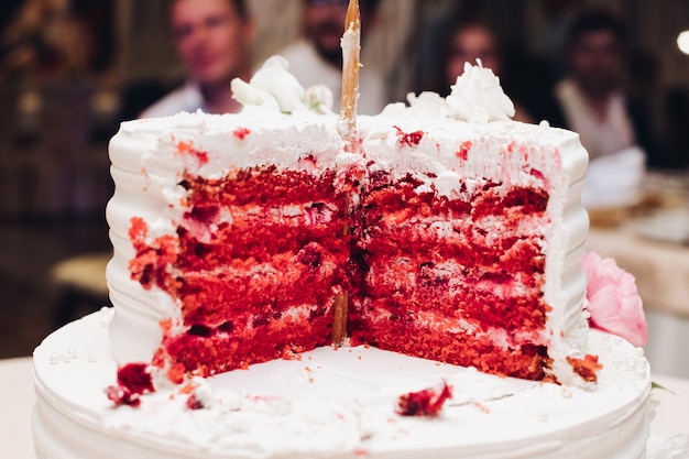 Close-up tasty big appetizing fresh piece of layered biscuit cake covered by white whipped cream icing. Beautiful dessert food serving for banquet event people guest at background. red velvet