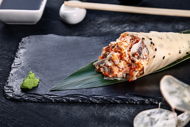 Close-up of tasty hand roll sushi in mamenori with eel and Tobico caviar served on dark stone plate with soy sauce and ginger