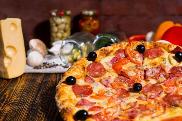 Close up of tasty pepperoni pizza with olives, near lie tomatoes, cheese, mushrooms and other vegetables on napkin in the background