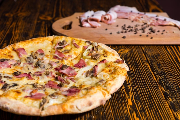 Primo piano di una gustosa pizza con varietà di condimenti e formaggio su un tavolo di legno, vicino a un tagliere con pancetta e spezie