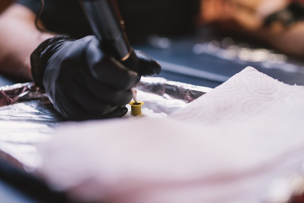 Photo close up of tattoo artists hand in black glove picking ink from ink bowl with a tattoo pen.