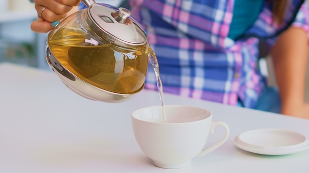 Close up of tea from the kettle slowly pour into porcelain cup. Young lady preparing green tea in the kitchen in the morning at breakfast, using teapot, teacup and healty herbal leaves.