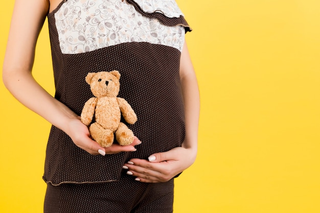 Close up of teddy bear in pregnant woman's hand at yellow background. Future mother is wearing brown pajamas. Motherhood concept. Copy space.