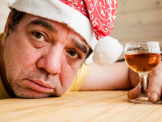 Foto close-up di un uomo maturo pensieroso che indossa il cappello di babbo natale con un bicchiere di alcol a casa