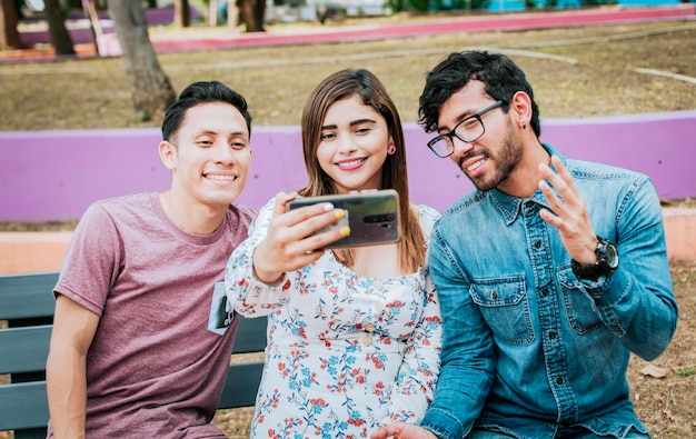 Close up of three friends taking a selfie sitting in the park Three smiling friends sitting in the park taking a selfie Front view of three happy friends taking a selfie while sitting on a bench