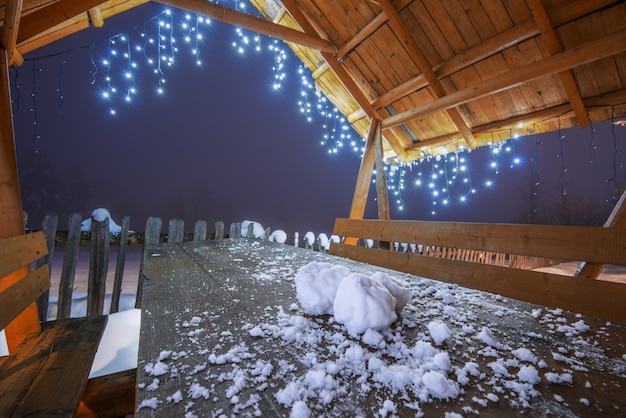 Close-up three snowballs lie on a wooden table under roof of arbor with New Year's lights. Picturesque mountainous place with spruce forest on frosty winter evening during Christmas holidays