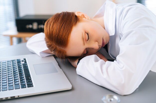 Close-up of tired overworked redhead female doctor in white coat sleeping at desk with laptop and stethoscope on workplace near window in office room of medic clinic.