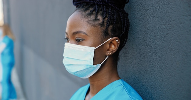 Close up stanco giovane donna afroamericana medico che toglie maschera medica e sorseggiando una bevanda calda mentre si riposa e si appoggia sulla parete all'aperto. infermiera femminile graziosa che beve caffè e riposo dopo il duro lavoro