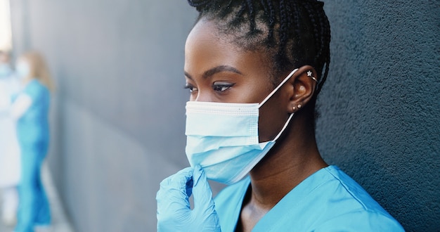 Close up tired young african american woman doctor taking off\
medical mask and sipping hot drink while resting and leaning on\
wall outdoor. pretty female nurse drinking coffee and rest after\
hard work