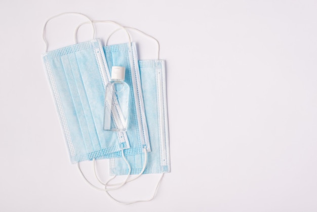 Close up top above overhead view photo of three medical blue masks and bottle with sanitizing liquid lying on white table with copy space