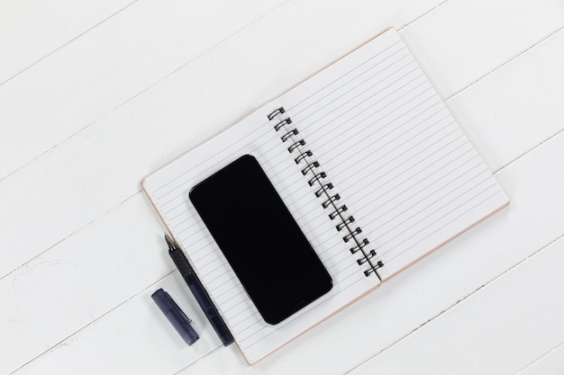 Close up top view of a black smartphone, a notebook and a black pen arranged on a plain white background