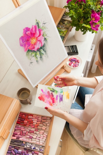 Close-up top view girl artist chooses dry pastel colors for drawing of pink rosehip flower sitting at desk with palette and easel and a bouquet of rosehips. Creativity and hobby concept