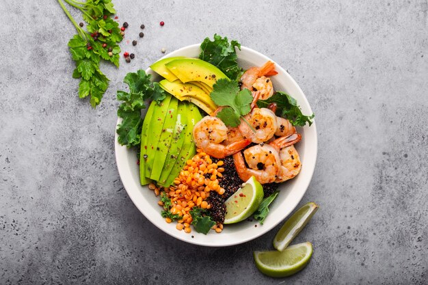 Close-up, top view of salad bowl with shrimps, avocado, fresh kale, quinoa, red lentils, lime and olive oil on gray stone background. Lunch bowl, healthy clean eating, dieting or nutrition concept