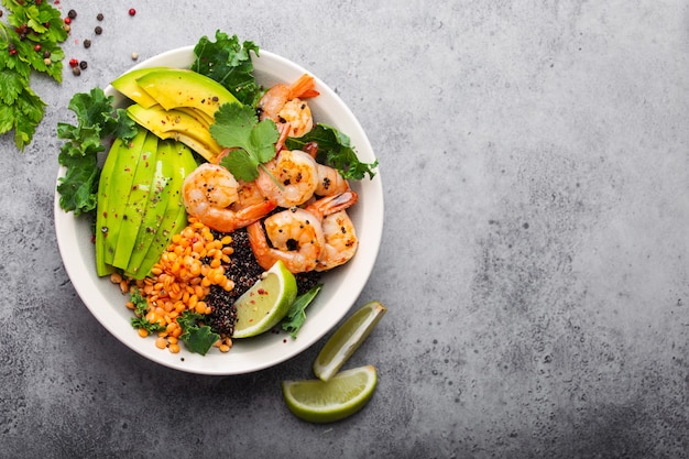 Close-up, top view of salad bowl with shrimps, avocado, fresh kale, quinoa, red lentils, lime and olive oil on gray stone background with space for text. Lunch bowl for healthy eating or dieting