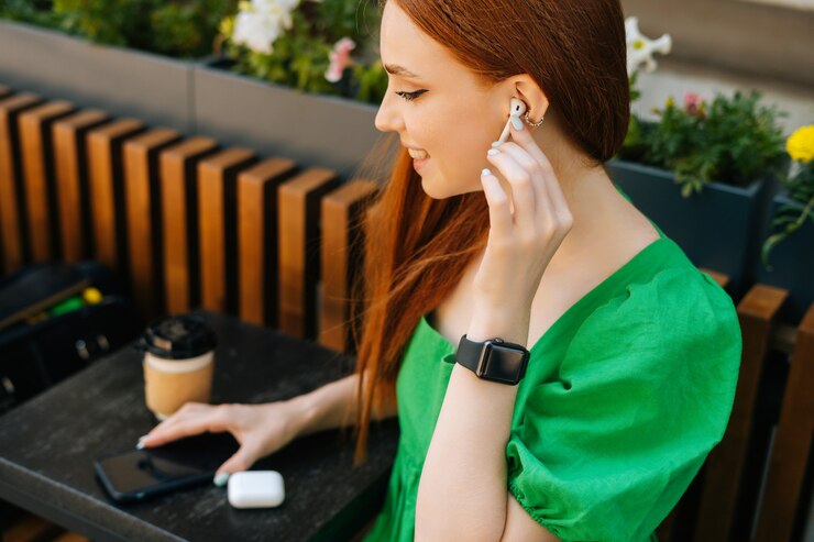 Photo close-up top view of smiling young woman touching wireless earphones, listening music using mobile phone sitting at table, in outdoor cafe terrace in summer day, blurred background, selective focus