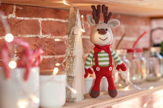 Close up toy amigurumi deer in striped sweater and stylish red butterfly tie stands on the wooden shelf near the decorated christmas tree and christmas lights.