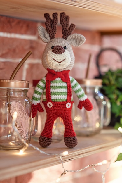 Close up toy amigurumi deer in striped sweater and stylish red butterfly tie stands on the wooden shelf near the decorated christmas tree and christmas lights.