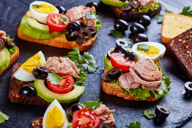 Close-up of tuna sandwiches with avocado slices, lettuce, tomatoes, black olives and hard boiled egg on rye and corn toasted bread slices on a black slate board with ingredients