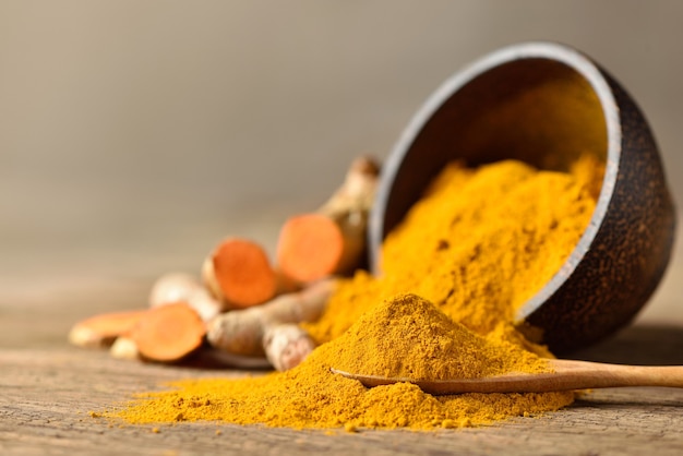 Close-up of Turmeric (curcumin) powder in  wooden spoon with bowl and fresh rhizome slices on wood background.