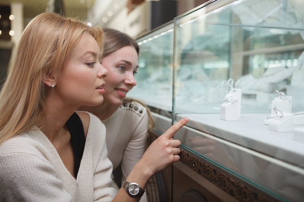 Photo close up of two beautiful women choosing jewelry to buy from retail display