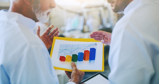 Close up of two business partners in sterile white uniform discussing about sale growth of food products. Selective focus on chart. Food plant interior.