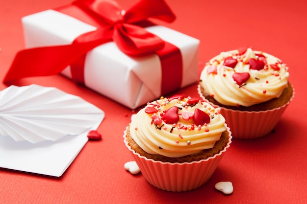 Close-up of two cupcakes with cream and heart decor on a red background with gift and envelope.