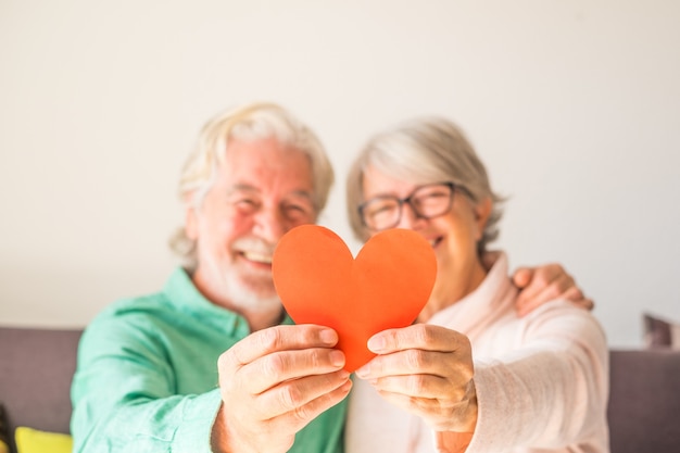 Primo piano di due anziani felici e innamorati che sorridono e guardano la telecamera tenendo insieme un cuore rosso - persone mature innamorate