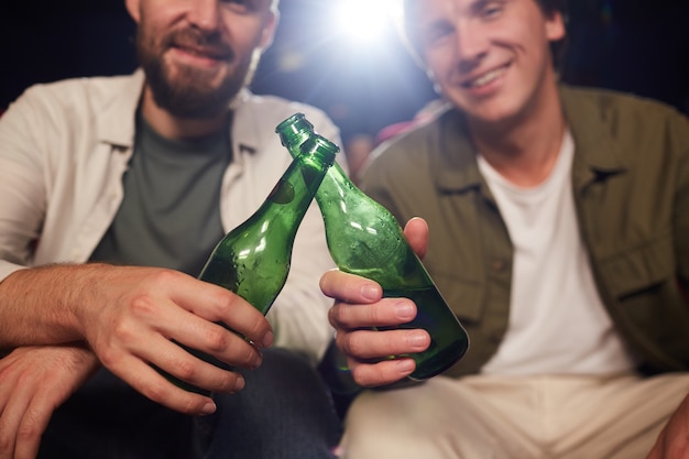 Close up of two male friends smiling and clinking beer bottles while watching movie in cinema theater, copy space