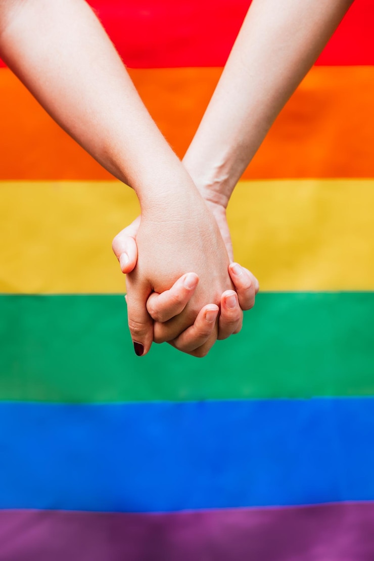 Photo close up of two womans hands holding each other with the lgbtq rainbow flag in the background