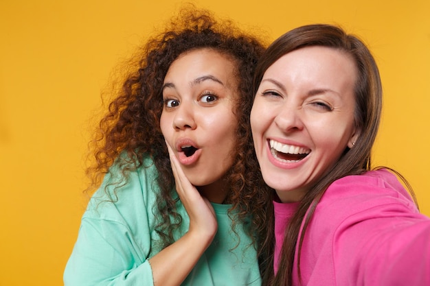 Foto primo piano di due donne amiche afroamericane europee in abiti rosa verdi in posa isolata su sfondo giallo. concetto di stile di vita delle persone. mock up spazio di copia. facendo selfie sul cellulare.