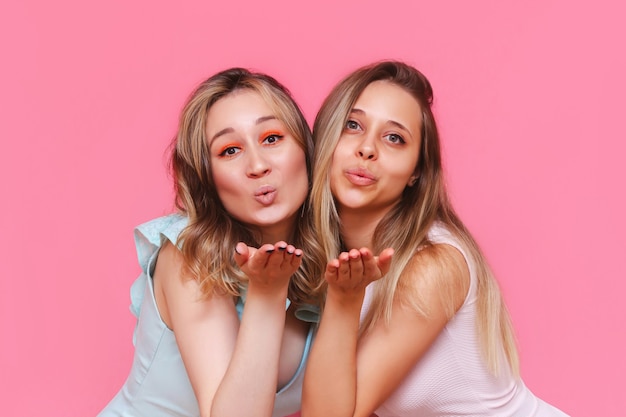 Close up of two young caucasian beautiful elegant women in stylish dresses send air kiss with open palms looking at front isolated on a light color pink wall